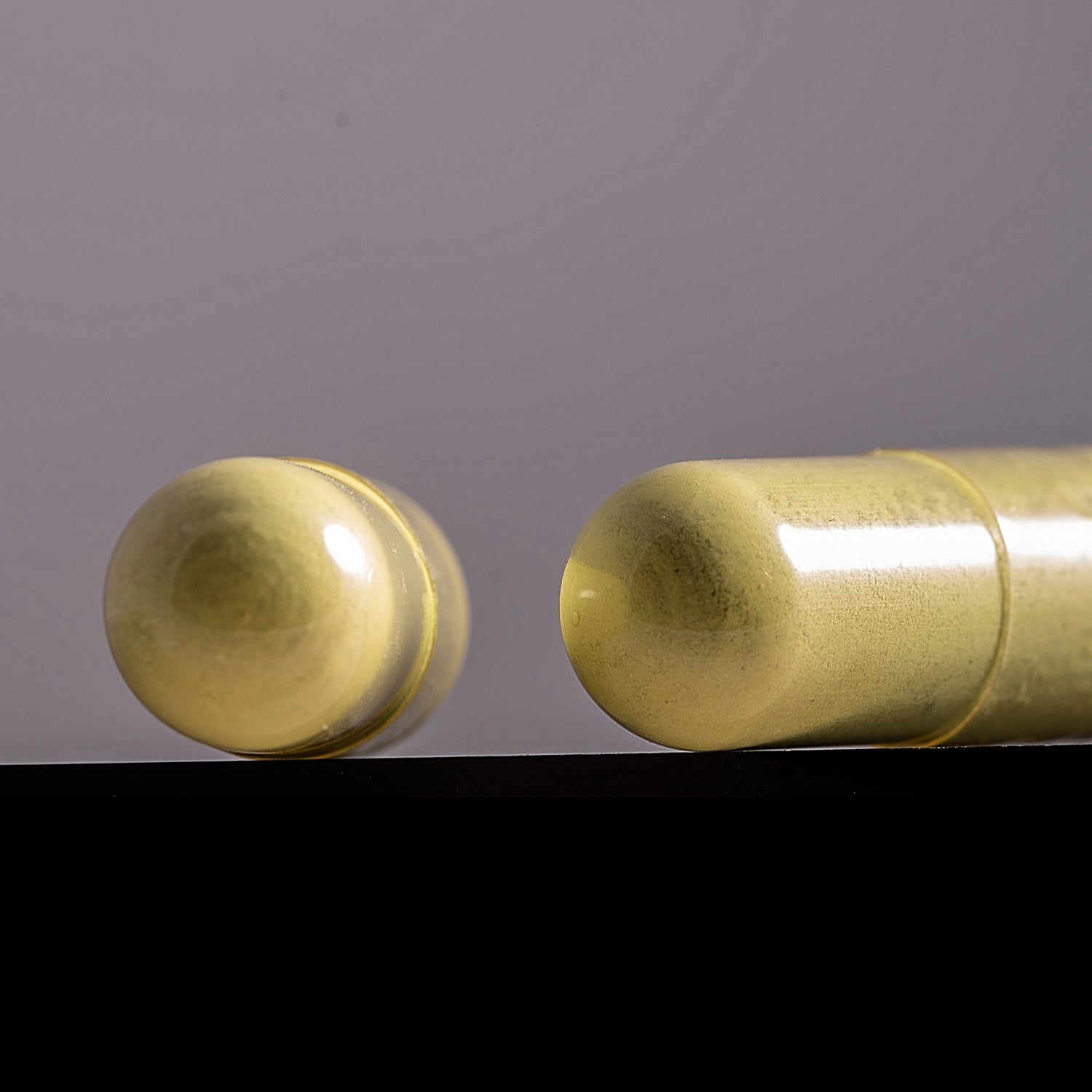 A close-up of a gold capsule-shaped cosmetic product, showing the open rounded dome on the left and the sealed capsule body on the right, resting on a black surface against a soft gray background.