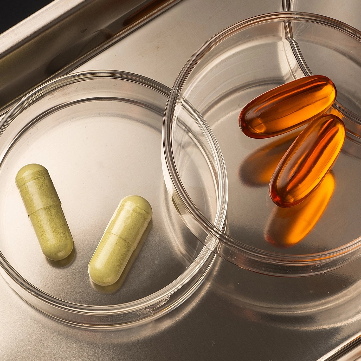 Two clear petri dishes on a metallic surface, one containing two pale yellow-green capsules and the other containing two translucent orange softgel capsules.