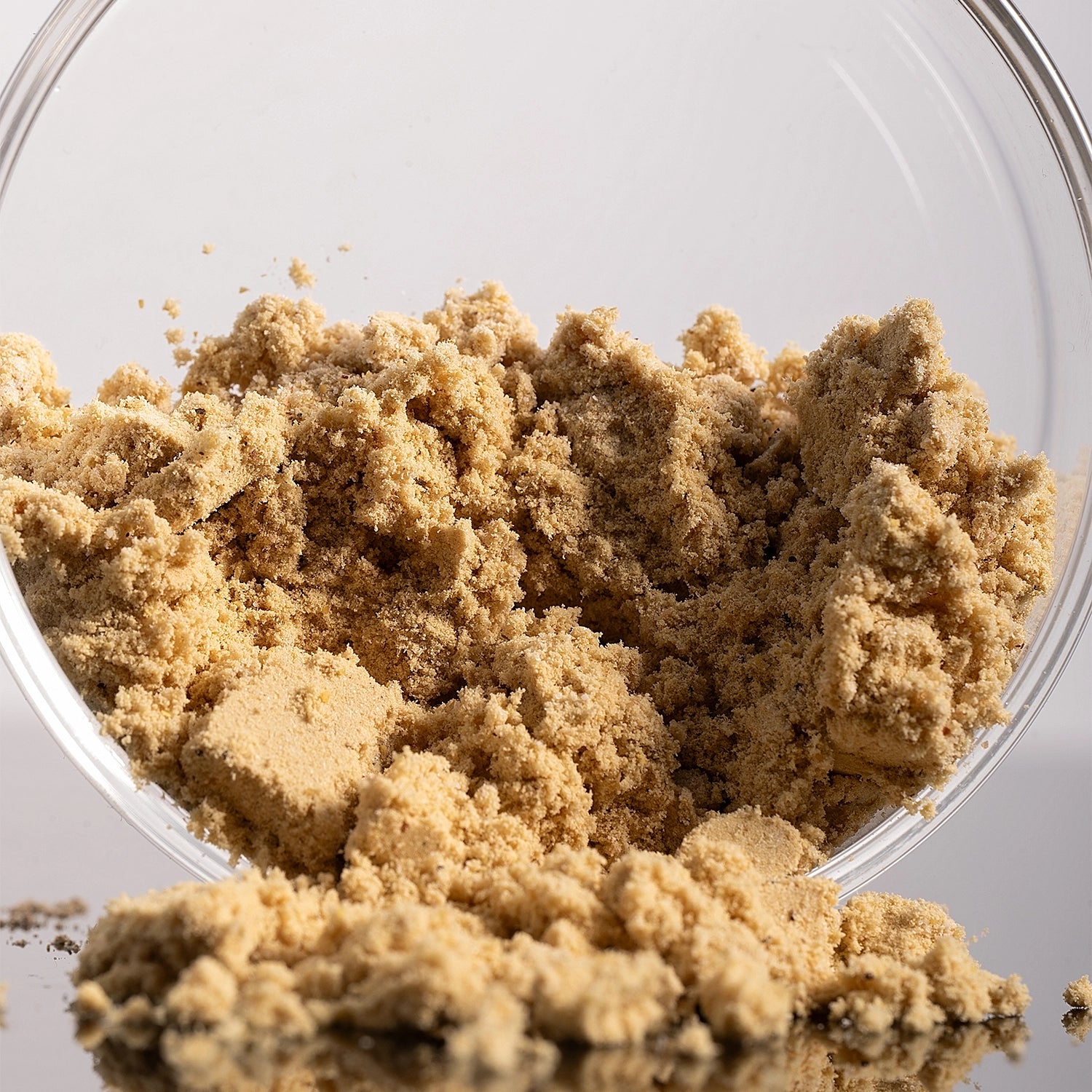 Light brown sugar granules spilling out of a clear glass bowl onto a reflective surface, photographed in a close-up studio setting.