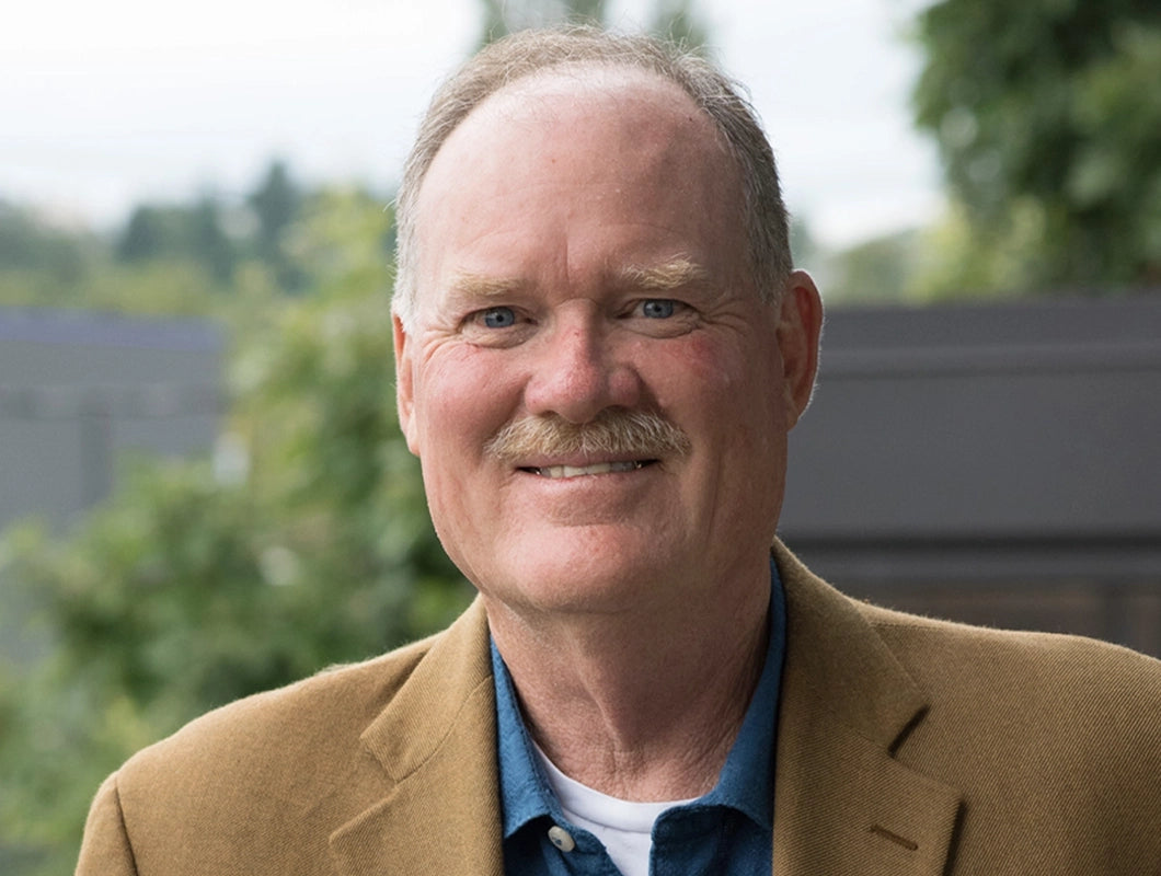 A smiling older man with a mustache, wearing a tan blazer over a blue button-up shirt and white undershirt, photographed outdoors with a blurred green and gray background.